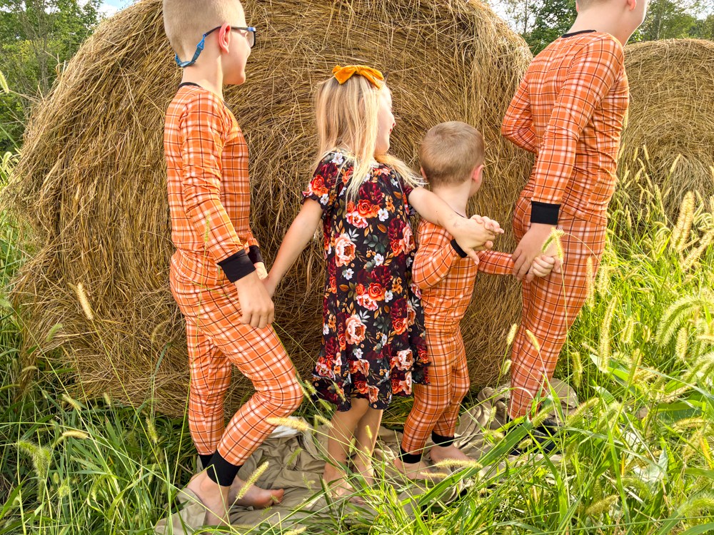 Four children standing outdoors in front of large hay bales, wearing matching orange plaid pajamas and floral nightgown.