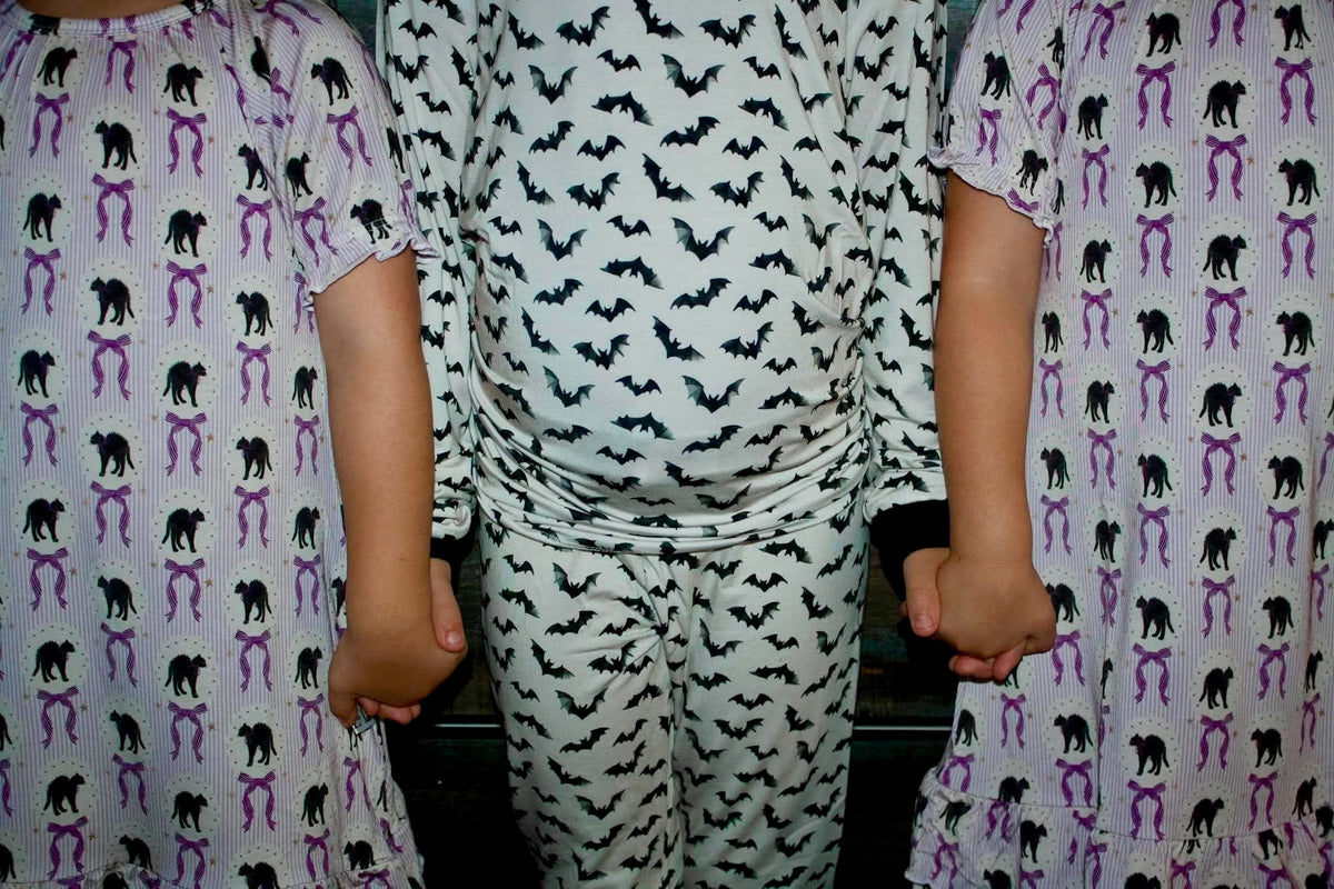 Three children wearing matching Halloween-themed pajamas, with the center child in a white long-sleeve top and pants set printed with black bats, flanked by two children in white pajamas with black cats and purple bows.