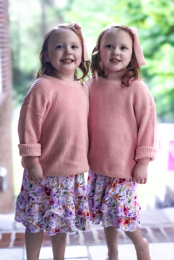 Two young girls standing side by side wearing matching pink ribbed knit sweaters and floral print nightgowns, with pink bows in their hair, set against a soft-focus outdoor background.