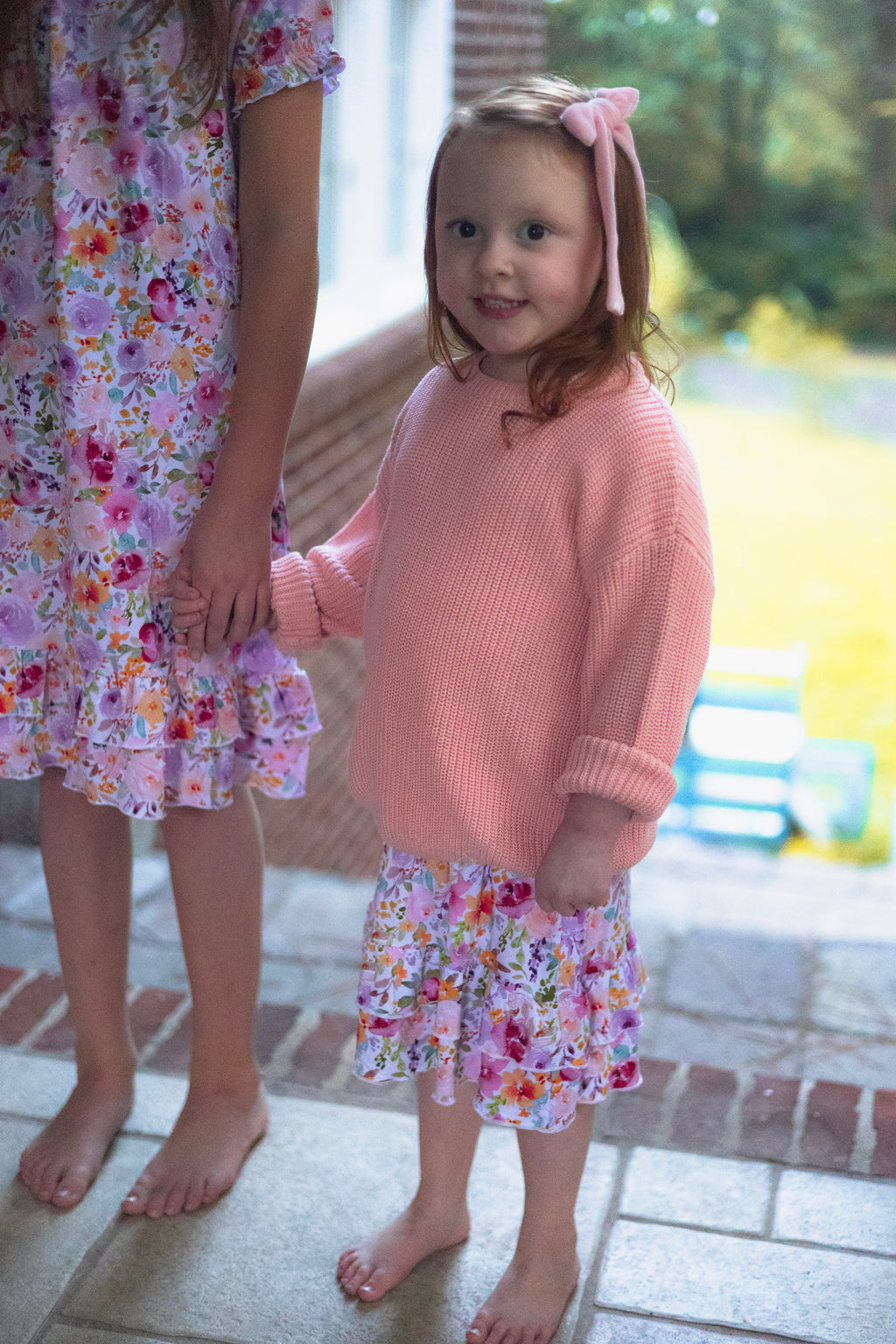 A young girl wearing a pink ribbed knit sweater and a pink floral nightgown, standing barefoot on a brick porch and holding hands with her older sister wearing a matching floral nightgown.