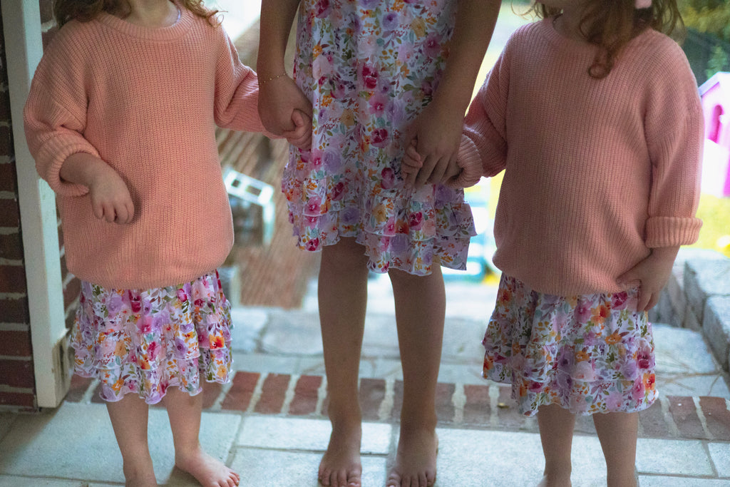 Three young girls standing barefoot on stone steps, wearing matching blush pink knit sweaters and floral print nightgowns.