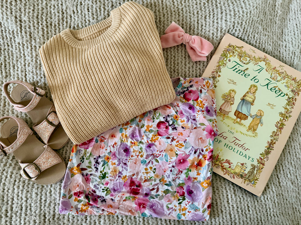 A styled flat-lay of a folded beige ribbed knit sweater, a folded pink floral skirt, a pink bow, glittery sandals, and a book arranged on a textured cream knit blanket.