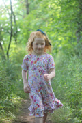 A young girl with curly red hair wearing a short-sleeve floral nightgown and a  bow, walking on a dirt path in a lush green forest.