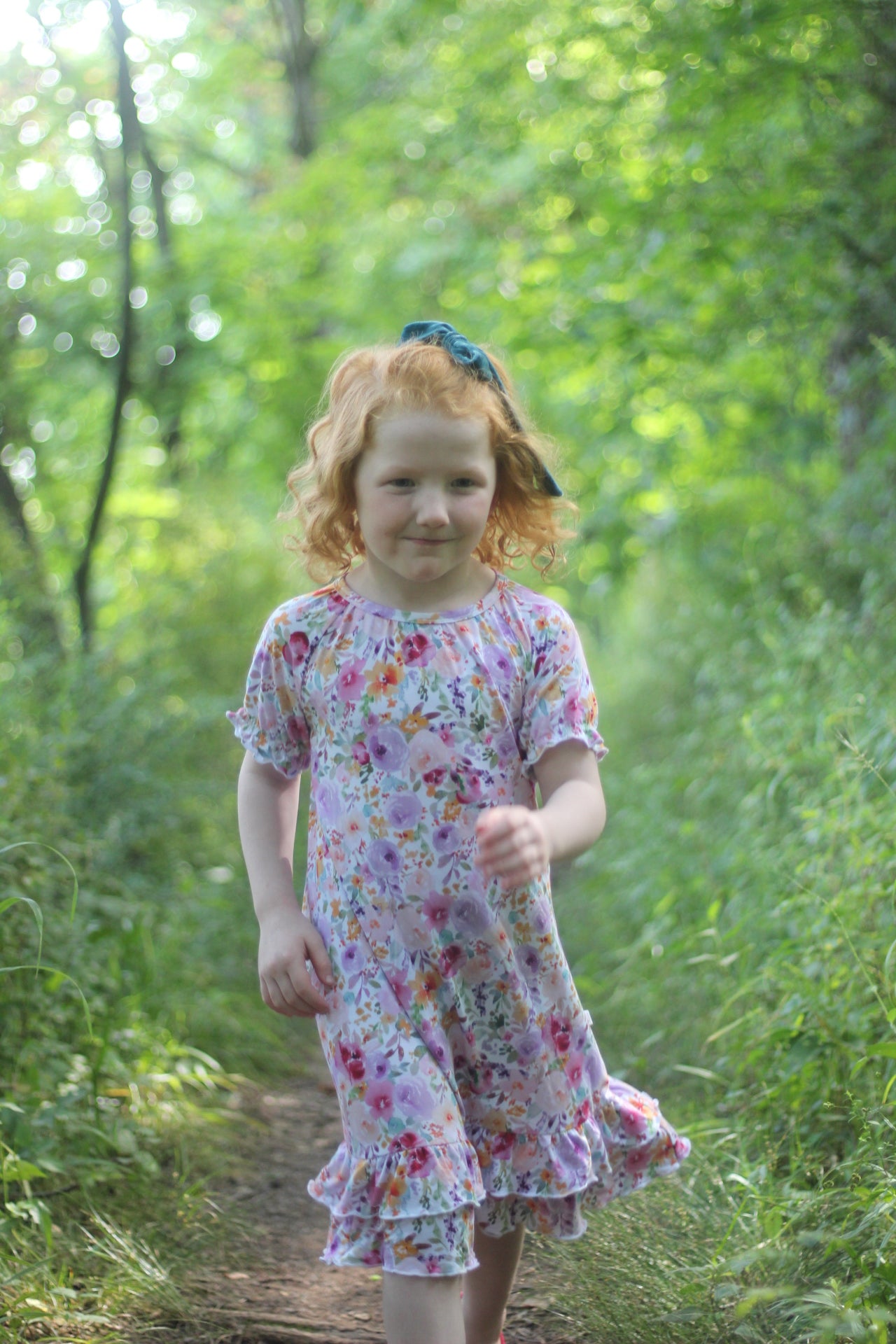 A young girl with curly red hair wearing a short-sleeve floral nightgown and a  bow, walking on a dirt path in a lush green forest.
