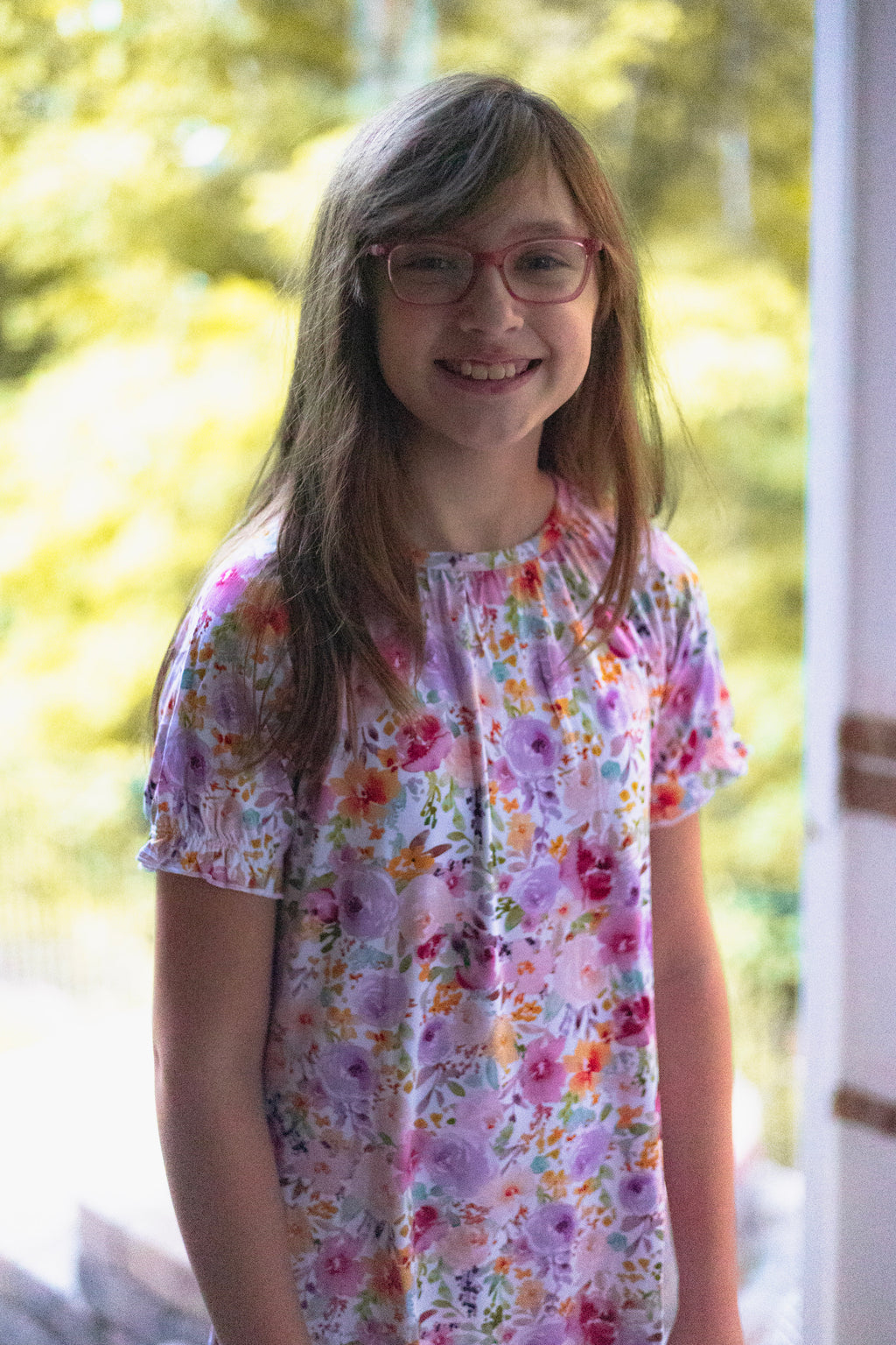 A young girl smiling, wearing a short-sleeve white blouse with a colorful floral print in pink, purple, orange, and green.