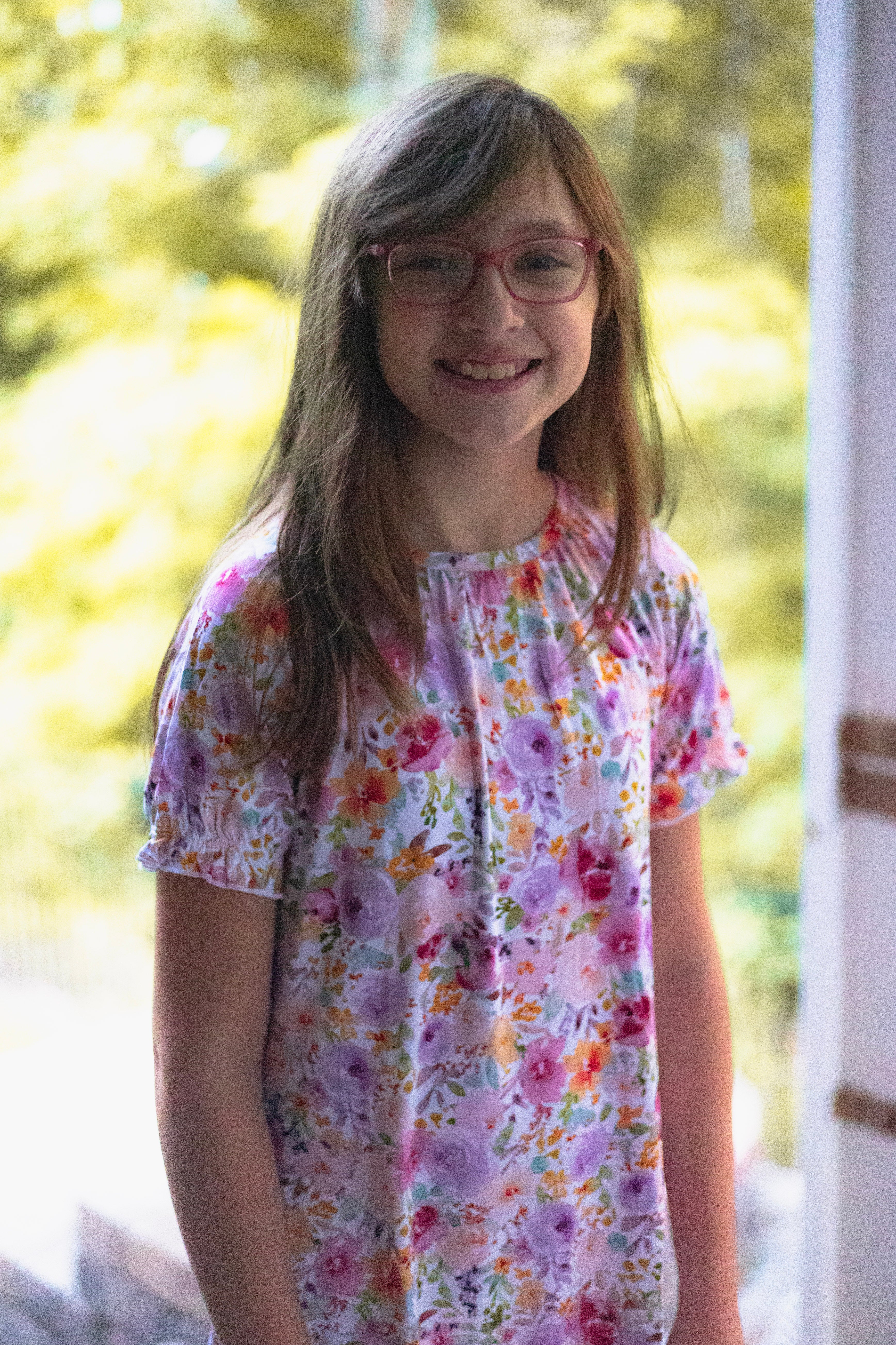 A young girl smiling, wearing a short-sleeve white blouse with a colorful floral print in pink, purple, orange, and green.