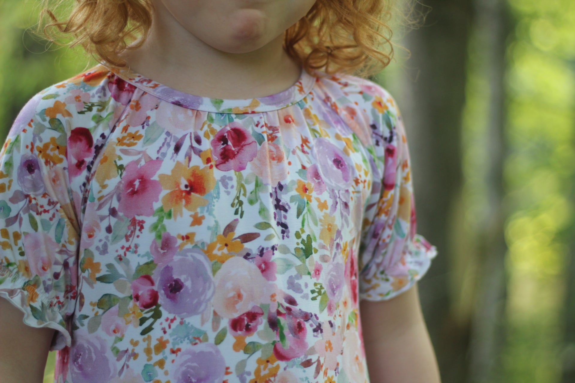 A close-up of a child wearing a short-sleeve top with a colorful watercolor floral print, set against a blurred green outdoor background.