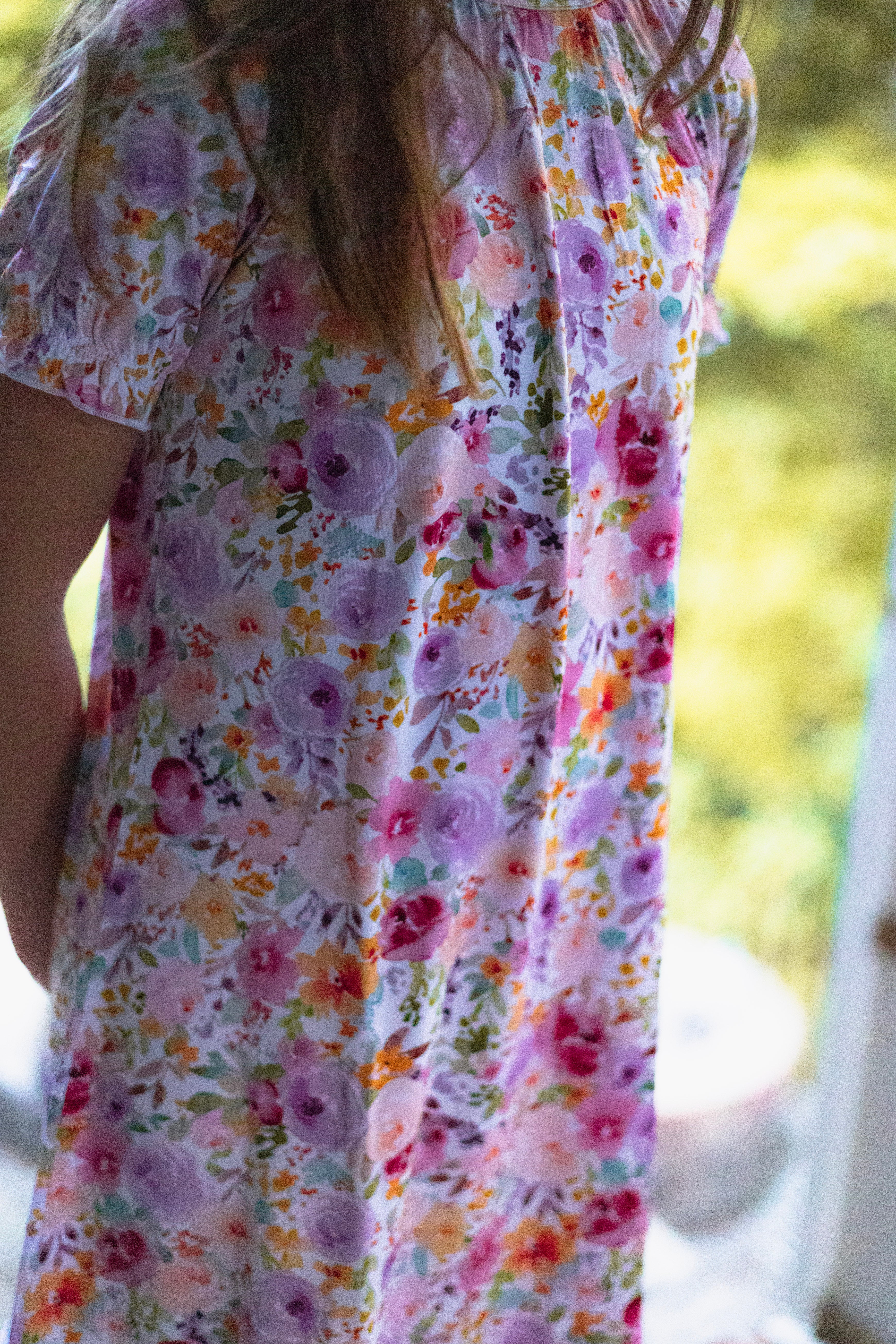 A close-up of a child wearing a short-sleeve nightgown with a colorful floral print in pink, purple, orange, and green on a white background.