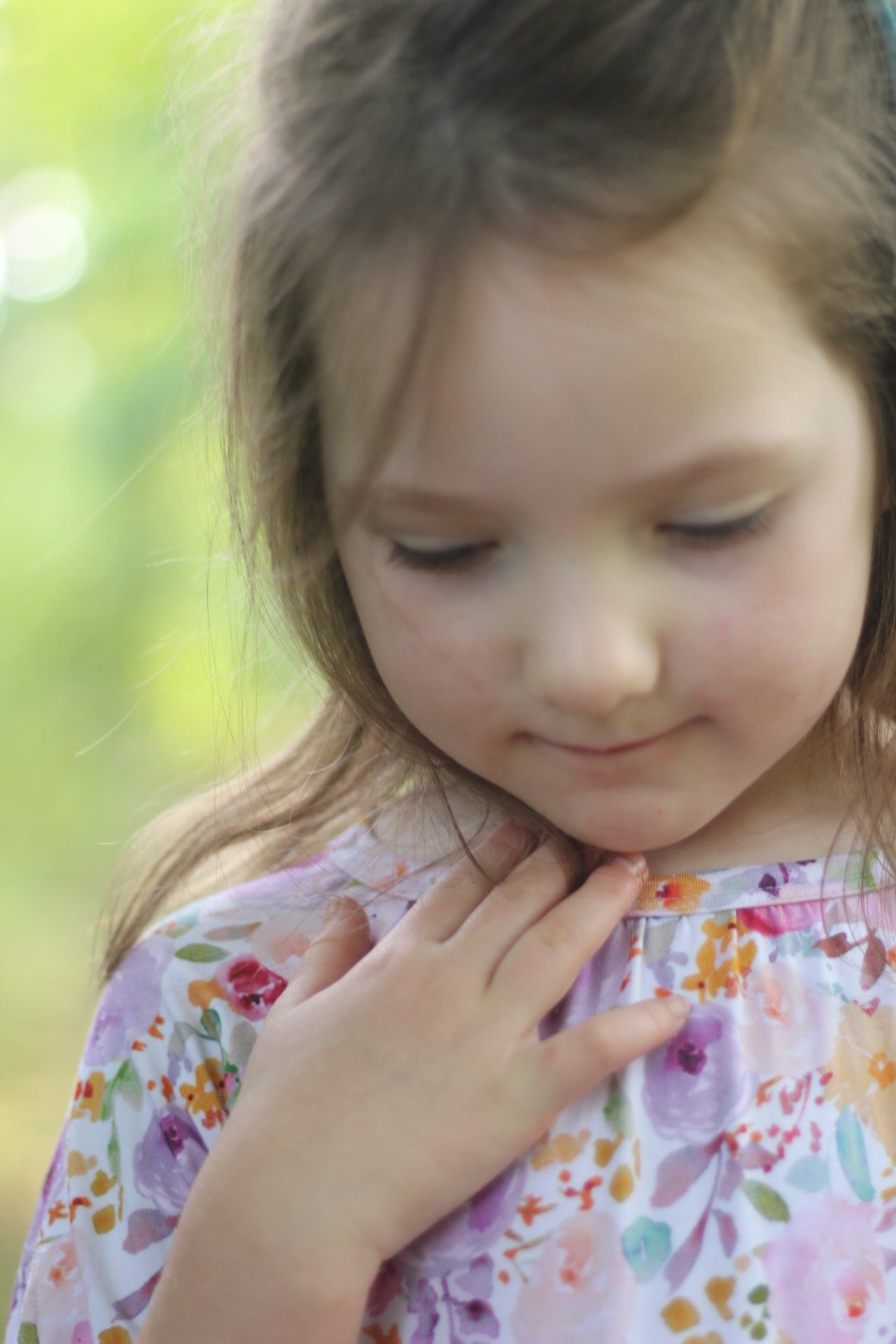 A close-up lifestyle shot of a young girl with light brown hair looking down, wearing a light purple floral print top with pink, orange, and green watercolor details.