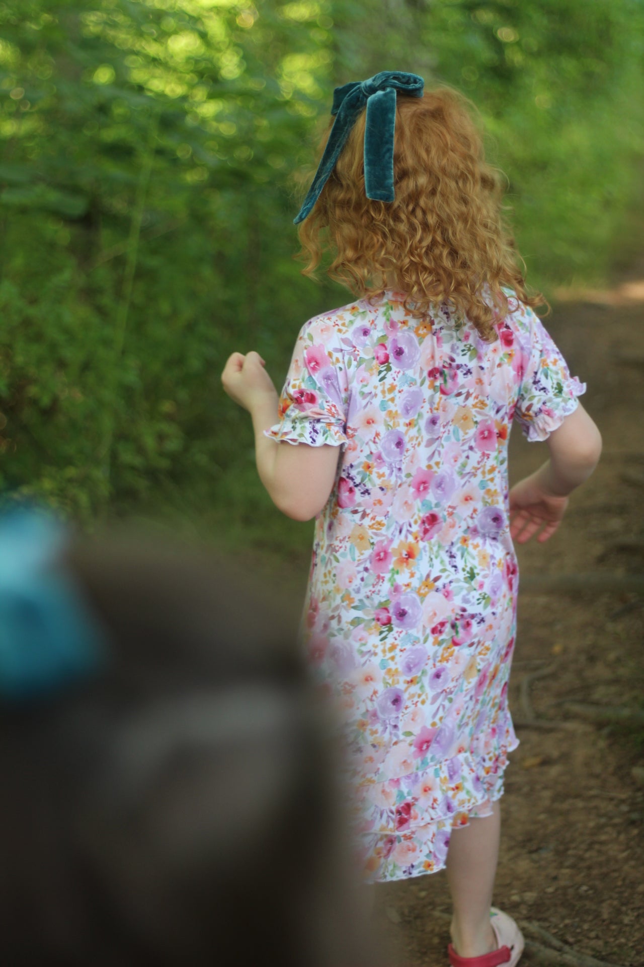 A child with curly red hair seen from behind, wearing a short-sleeve floral nightgown with a ruffled hem and a dark teal bow, walking on a forest path.