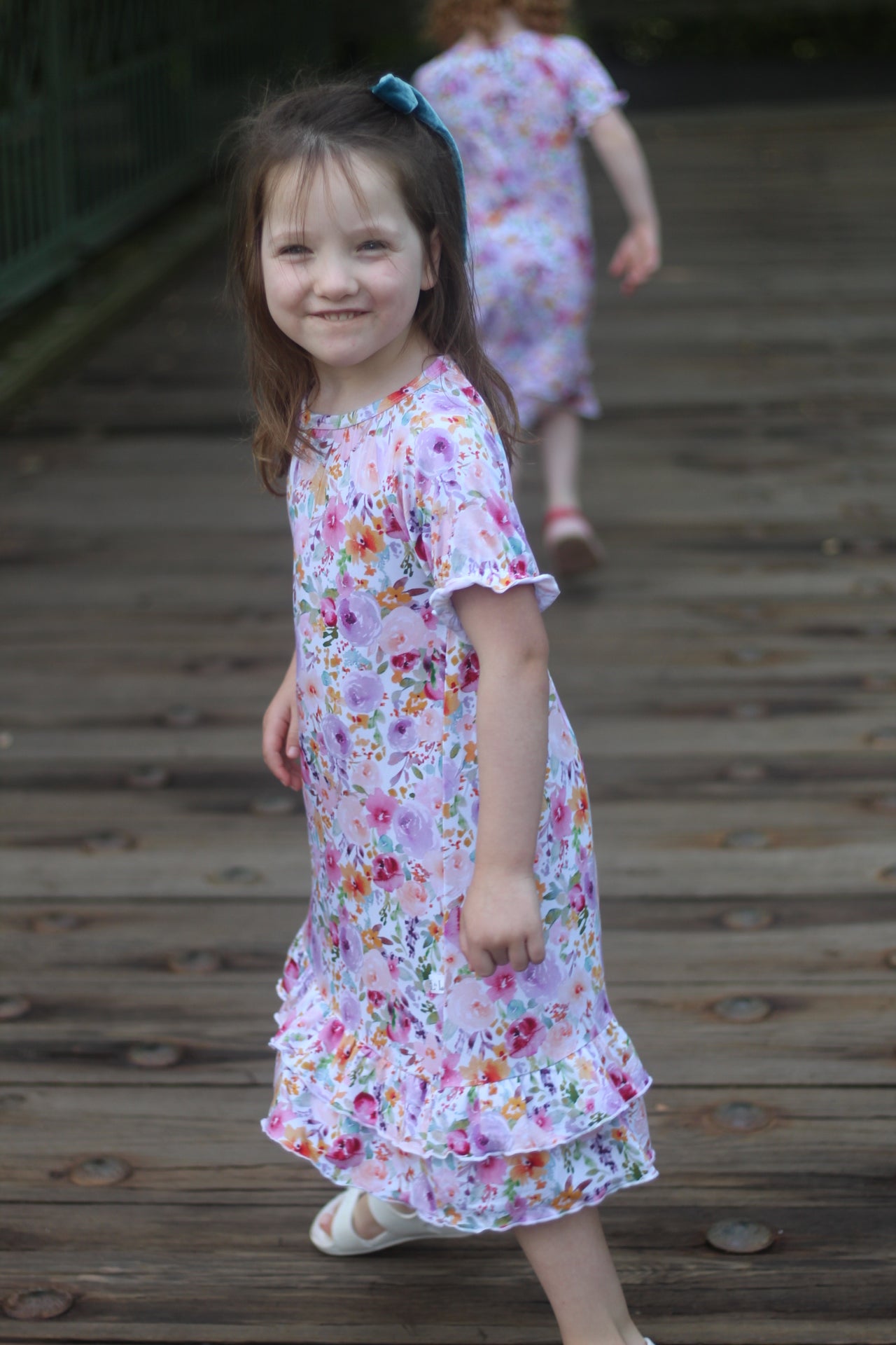 A young girl wearing a short-sleeve tiered floral nightgown walks on a wooden boardwalk, with another child in a matching dress blurred in the background.