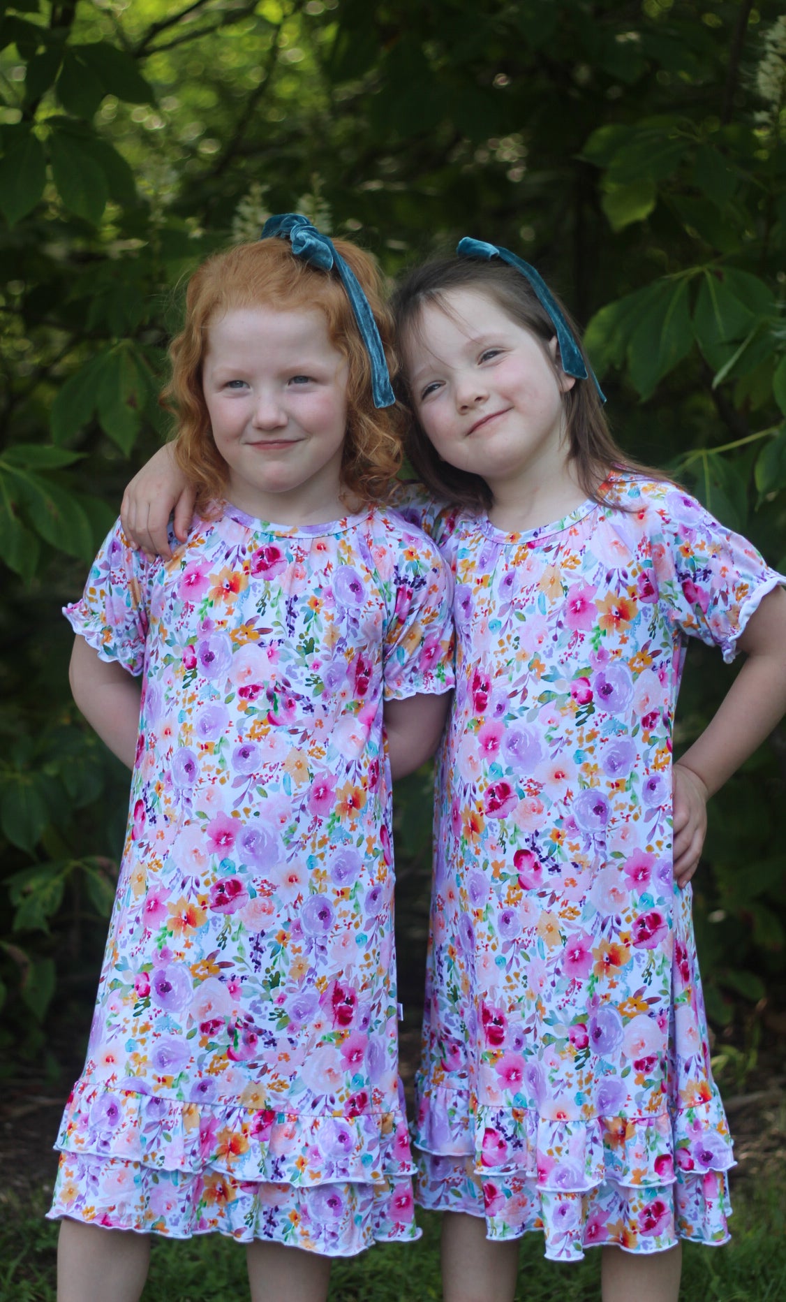 Two young girls wearing matching short-sleeve ruffled nightgowns with a colorful floral print on a light lavender background, standing outdoors in front of green foliage.