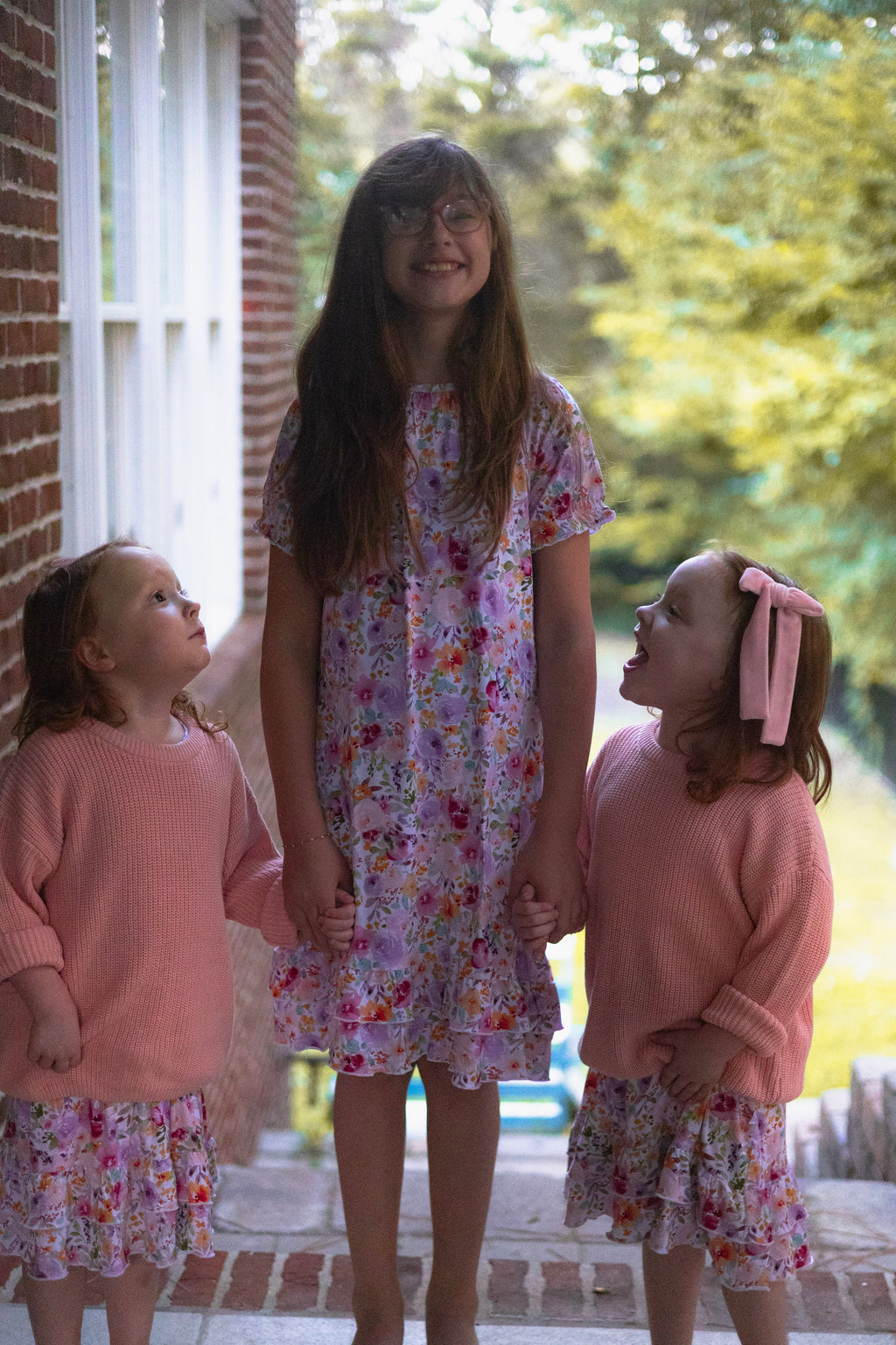 Three young girls standing together outdoors on a porch, wearing matching outfits. The center girl wears a pink floral dress, while the two girls on either side wear pink knit sweaters and matching nightgowns underneath.