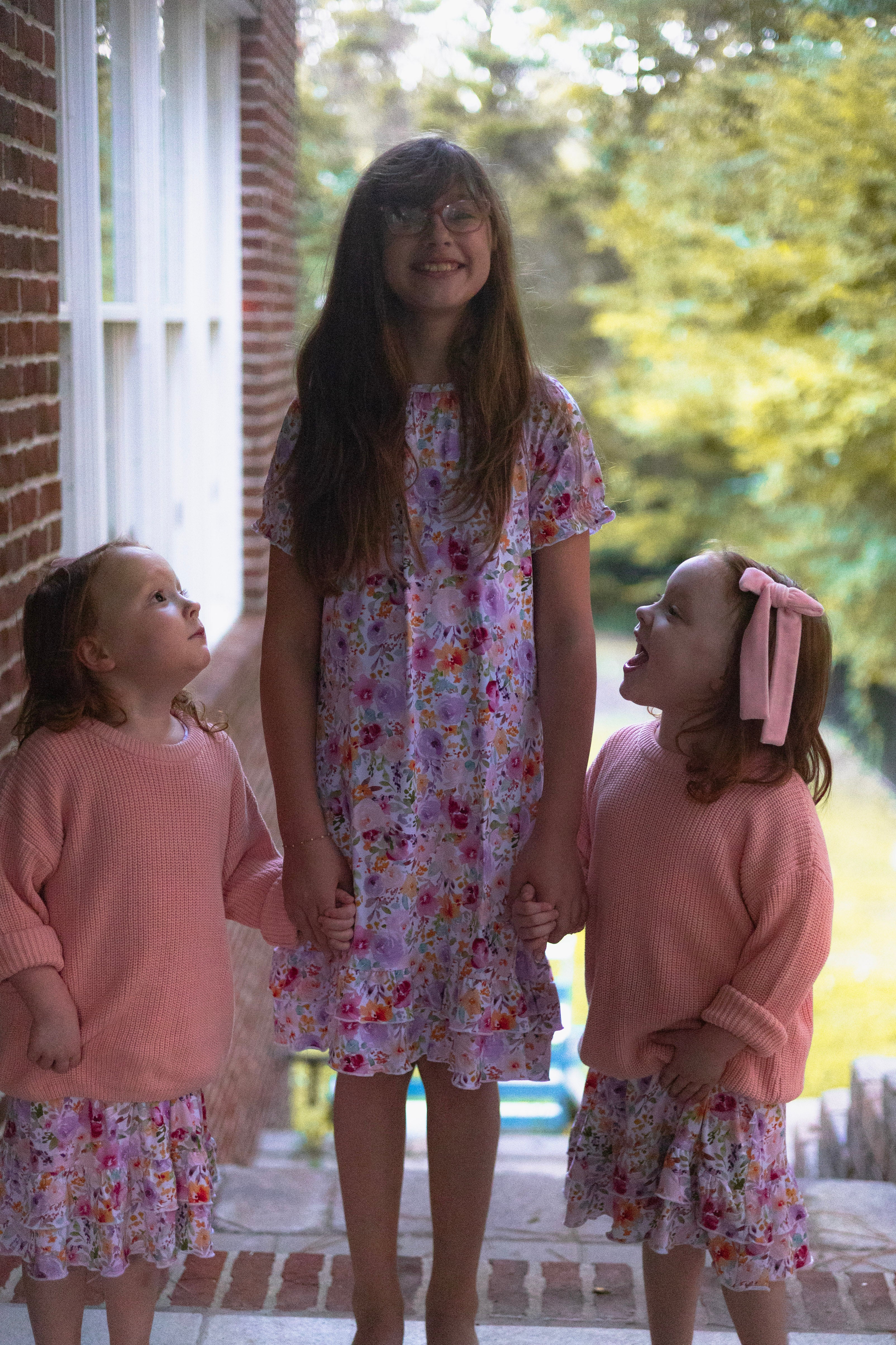 Three young girls standing together outdoors on a porch, wearing matching outfits. The center girl wears a pink floral dress, while the two girls on either side wear pink knit sweaters and matching nightgowns underneath.