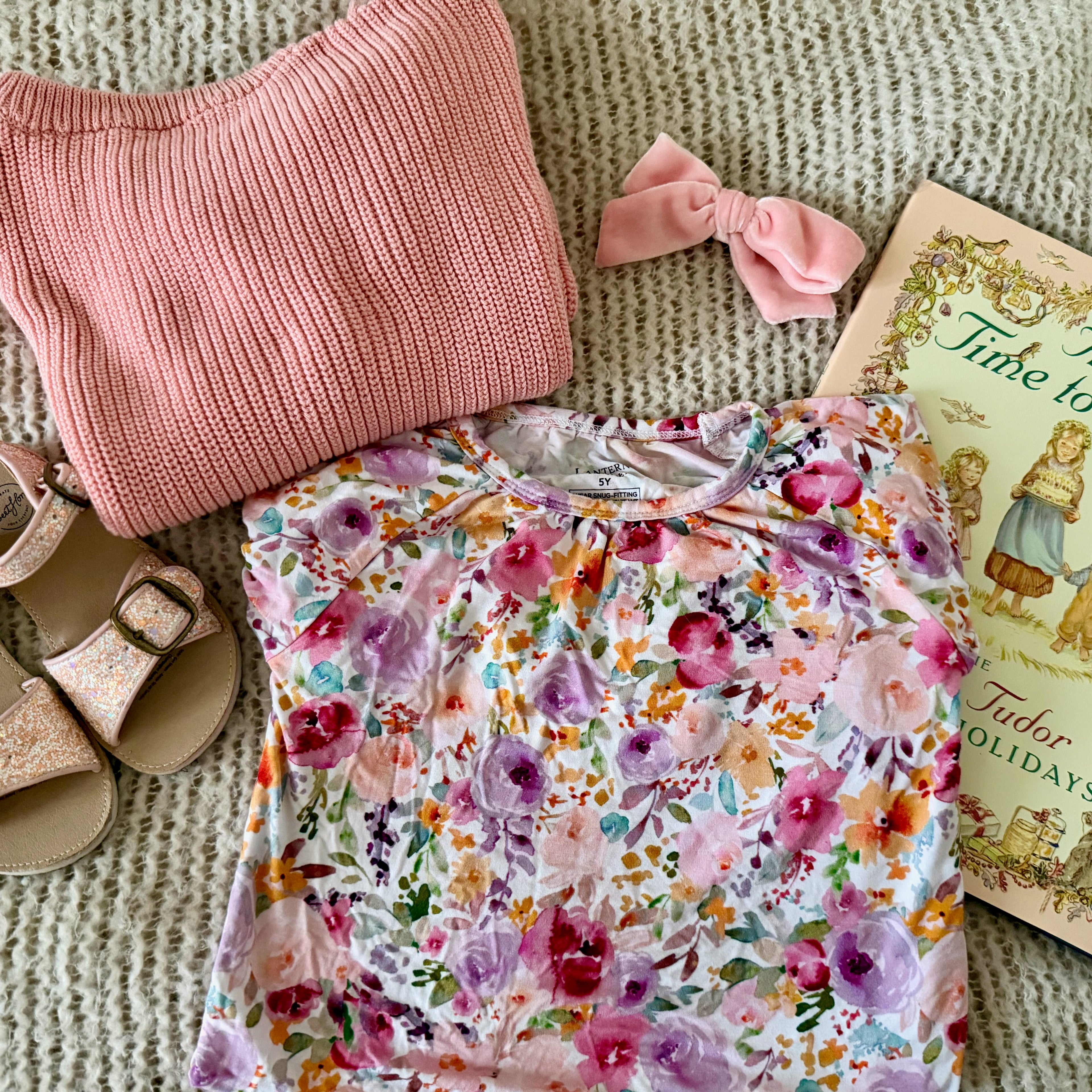 A styled flat-lay of a children's outfit featuring a pink ribbed knit sweater, a folded nightgown with a colorful floral print, a pink bow, and pink glitter sandals, arranged on a textured beige knit blanket next to a vintage book.