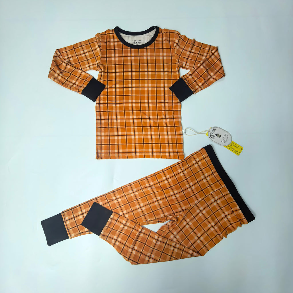 A two-piece children's pajama set laid flat on a light blue background, featuring a long-sleeve top and matching pants in an orange and yellow plaid pattern with black cuffs and collar trim. A product tag is attached to the shirt.