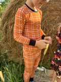 A child wearing a bright orange plaid long-sleeve pajama set with black trim, standing outdoors in front of a large hay bale and holding a wheat stalk.