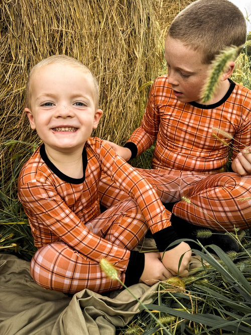 Two young boys sitting outdoors in a field of wheat, wearing matching orange and white plaid long-sleeve pajamas with black trim.