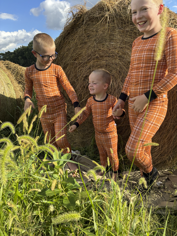 Three children wearing matching orange plaid long-sleeve pajamas stand outdoors in front of hay bales under a blue sky with clouds.