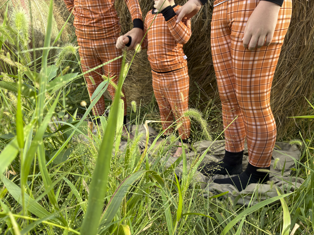 Three people wearing matching orange and white plaid long-sleeve pajamas, standing outdoors in tall green grass near a hay bale.