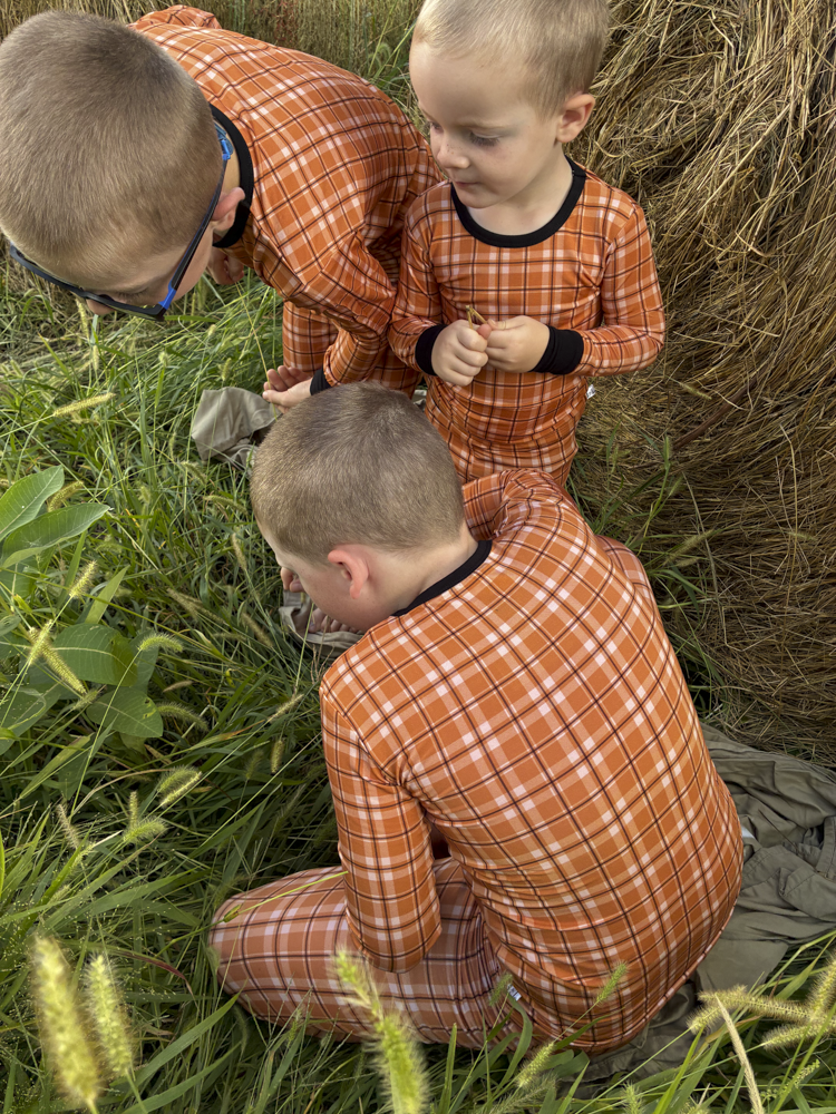 Three children wearing matching orange and white plaid long-sleeve pajamas with black trim, gathered outdoors in tall grass next to a bale of hay.