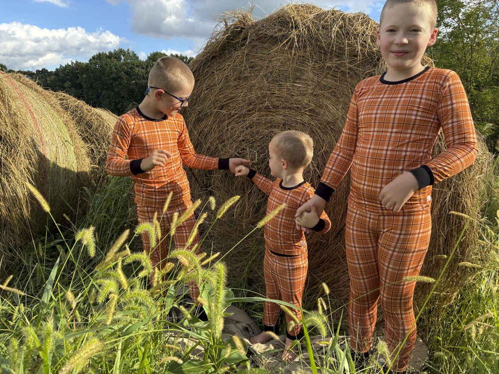 Three children wearing matching orange and brown plaid long-sleeve pajamas stand outdoors in front of hay bales under a blue sky.