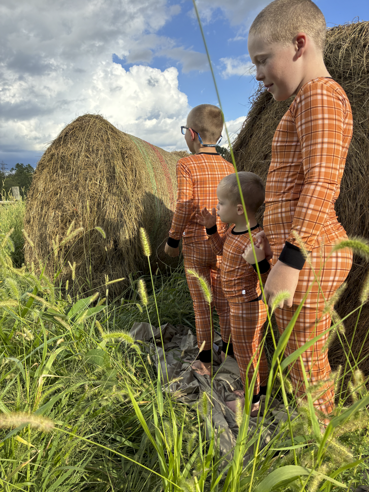 Three children wearing matching orange plaid pajamas stand outdoors in tall grass next to large hay bales under a blue sky with clouds.