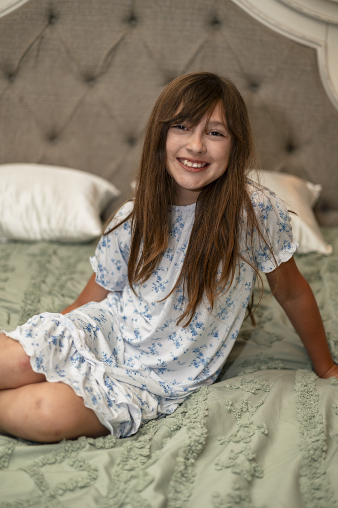 A young girl sits smiling on a bed wearing a white short-sleeve bamboo nightgown with a blue floral pattern.