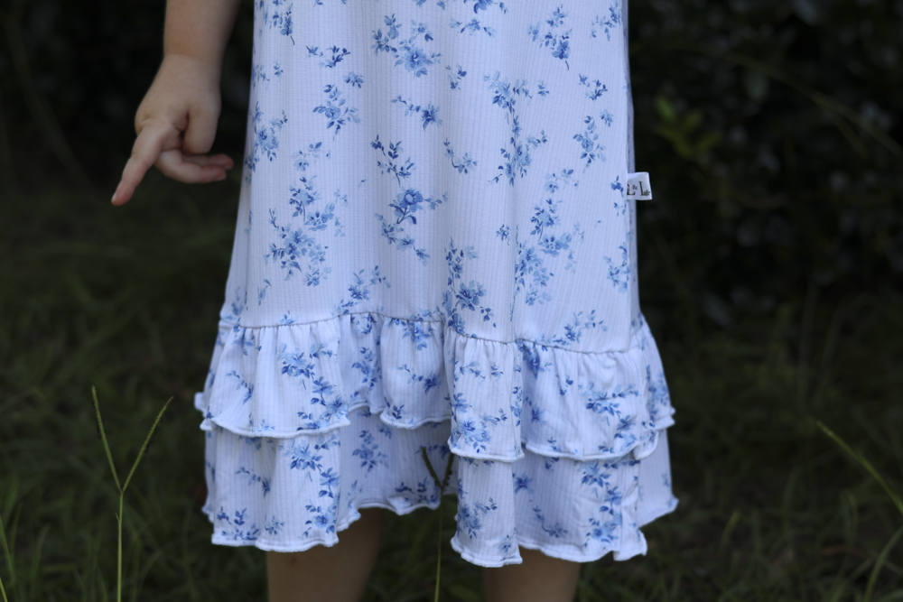 A close-up of a child wearing a white ruffled bamboo nightgown with a blue floral print, standing outdoors in a grassy area.