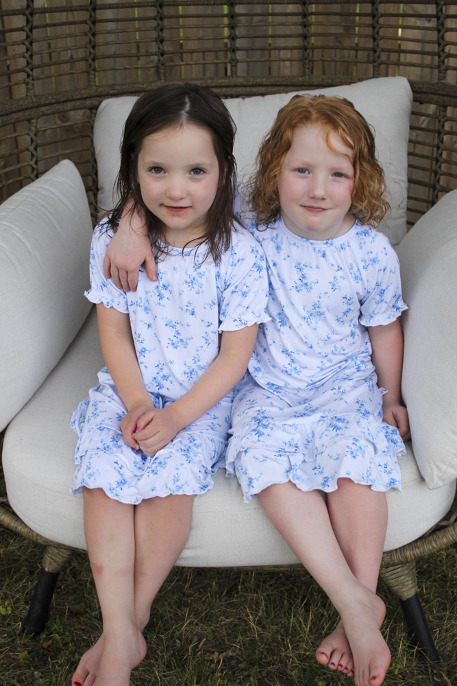 Two young girls sitting side-by-side on a white cushioned wicker patio chair, wearing matching light blue and white floral print short-sleeve bamboo nightgowns.