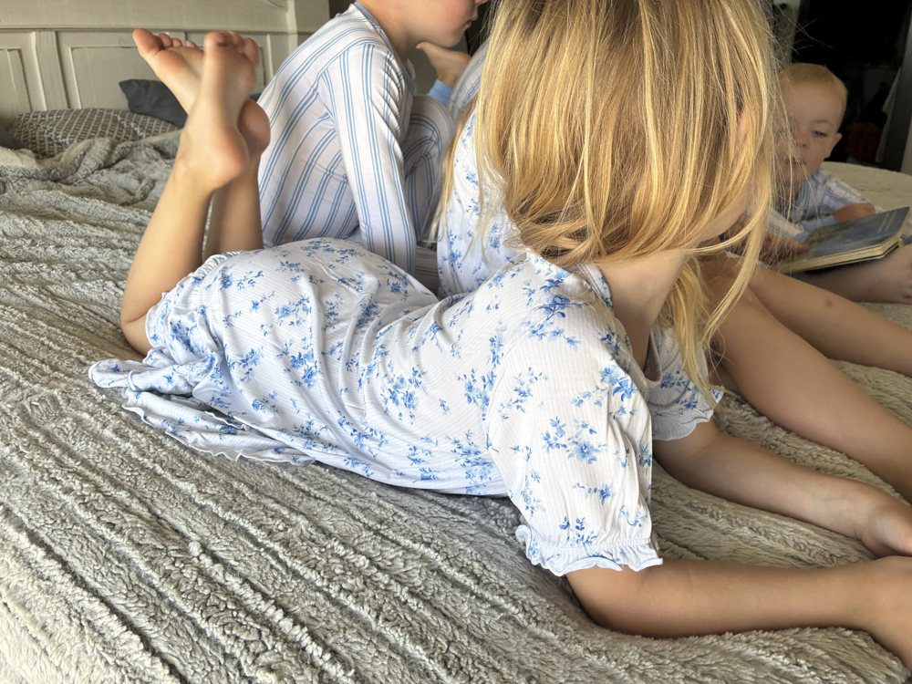 A young girl with blonde hair lies on a textured gray blanket wearing a white short-sleeve bamboo nightgown with a blue floral pattern. Two other children in matching bamboo pajamas are visible in the background on a bed.