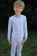 A young boy wearing a two-piece long-sleeve bamboo pajama set with light blue and white vertical stripes, standing outdoors against a dark green background of foliage and grass.