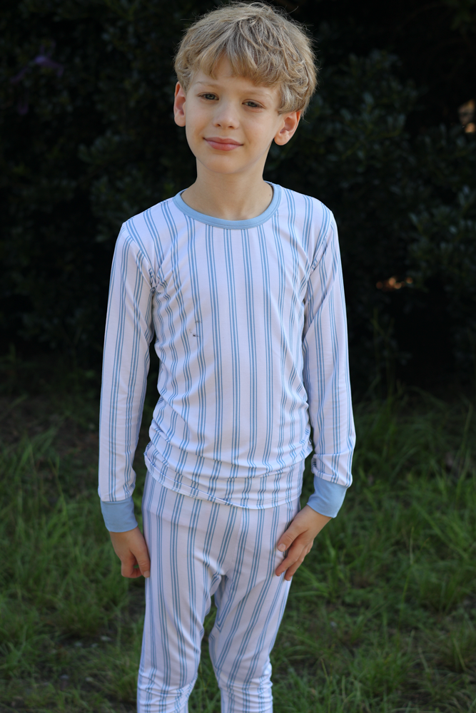 A young boy wearing a two-piece long-sleeve bamboo pajama set with light blue and white vertical stripes, standing outdoors against a dark green background of foliage and grass.