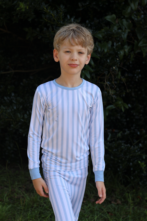 A young boy wearing a light blue and white vertically striped long-sleeve bamboo pajama set, standing outdoors against a dark green leafy background.