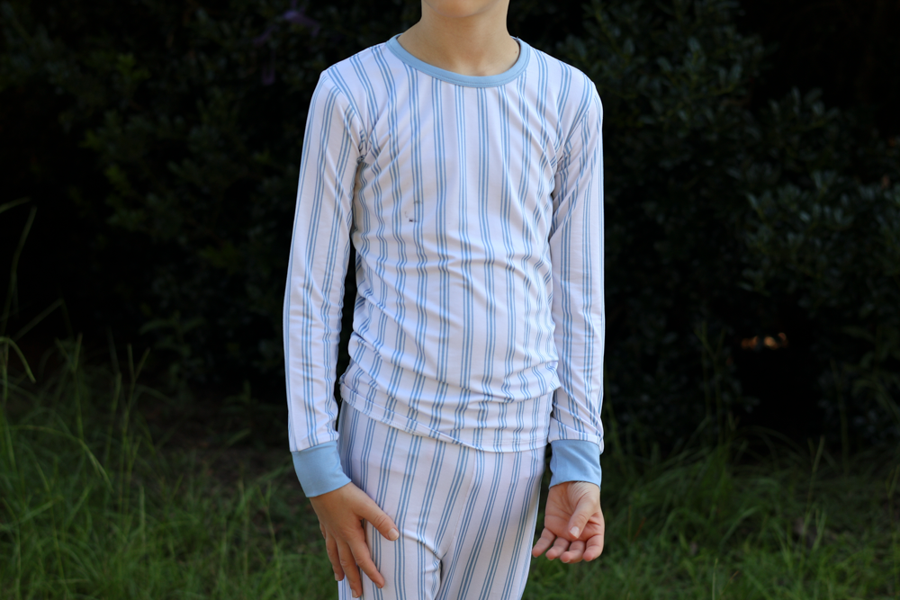 A person wearing a long-sleeve bamboo pajama top and matching pants with vertical light blue and white stripes, standing outdoors against a dark green foliage background.