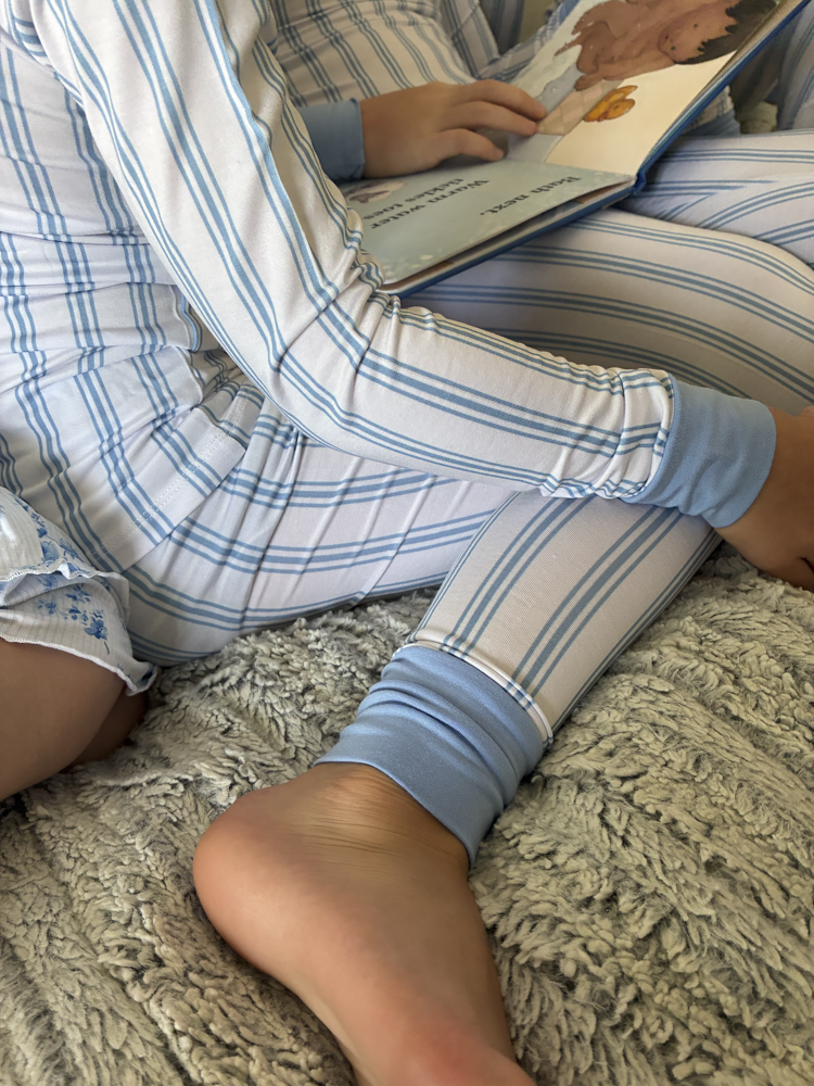 A person wearing light blue and white striped bamboo pajamas sits on a textured gray blanket, holding and reading a book.