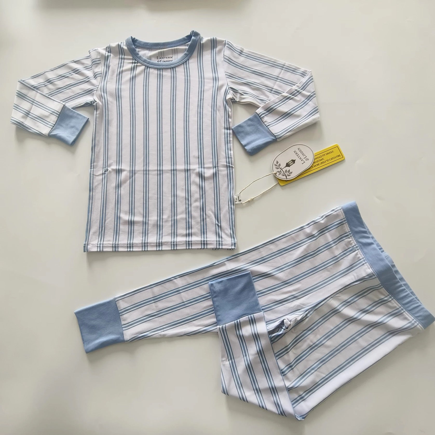 A two-piece children's bamboo pajama set laid flat on a light beige background. The set includes a long-sleeve top and matching pants, both featuring a white base with thin blue vertical stripes and solid light blue cuffs and pocket panels.