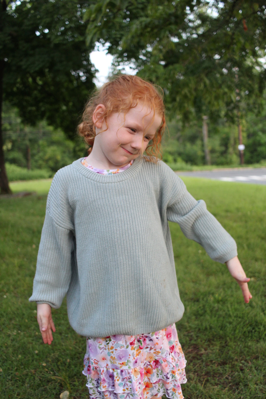 A young child wearing a sage green ribbed knit sweater and a colorful floral nightgown, standing outdoors on a grassy lawn with trees in the background.