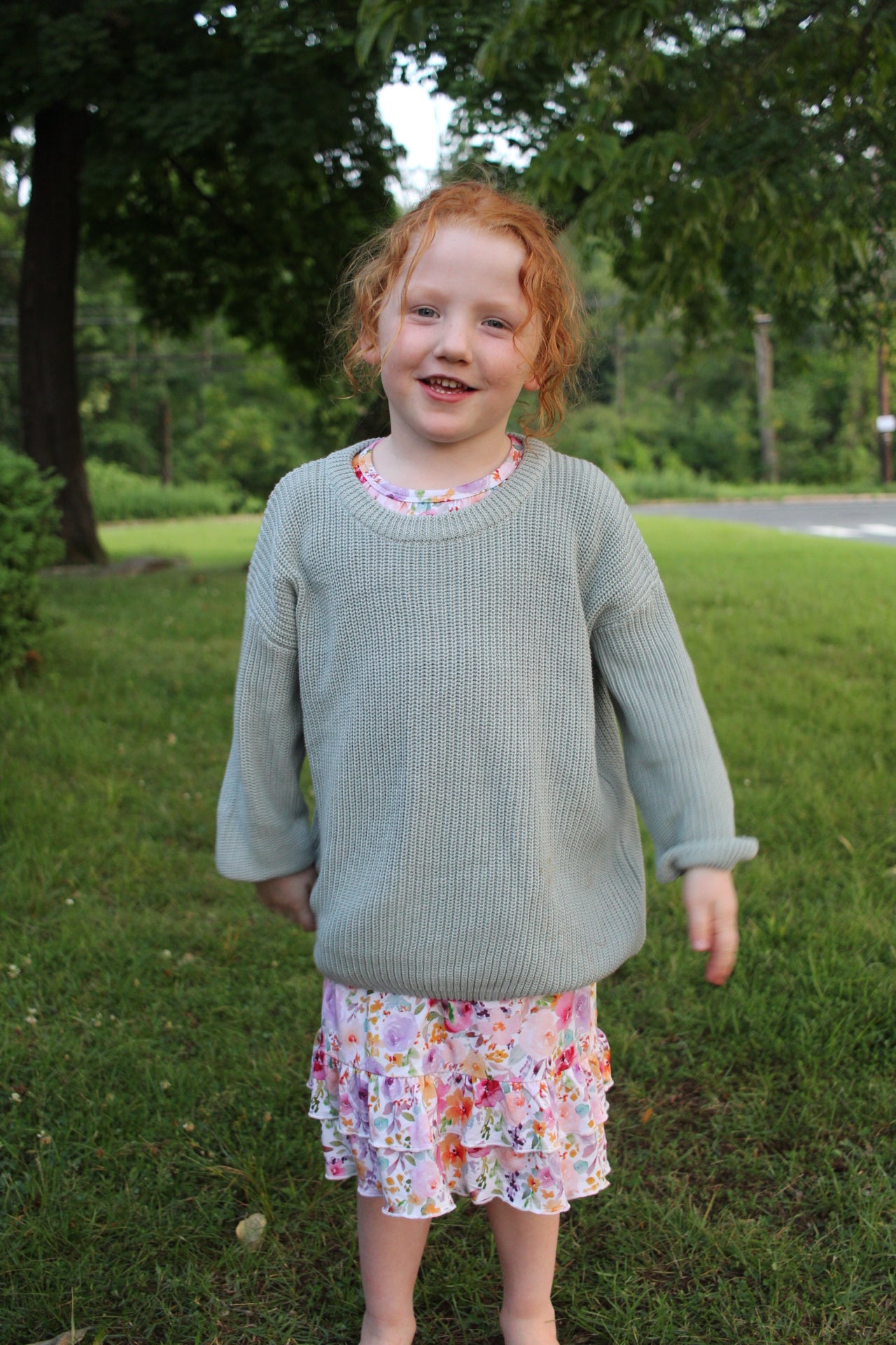 A young girl with red hair stands outdoors on a grassy lawn wearing a light sage green ribbed knit sweater and a floral nightgown.