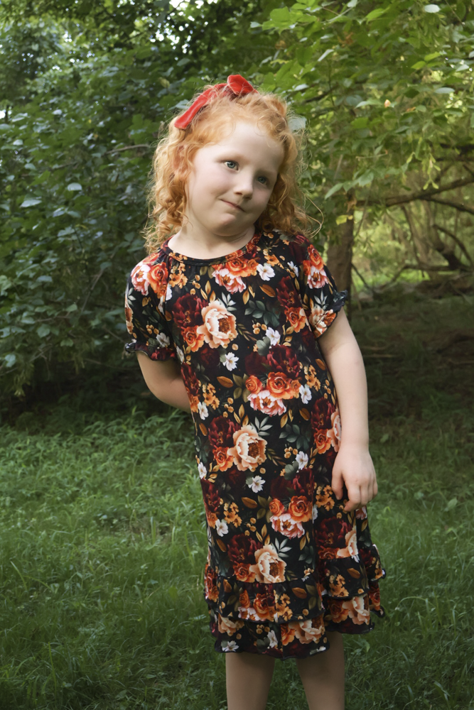 A young girl with curly red hair wearing a short-sleeve black nightgown with a vibrant orange, red, and white floral pattern, standing outdoors in a grassy, wooded area.