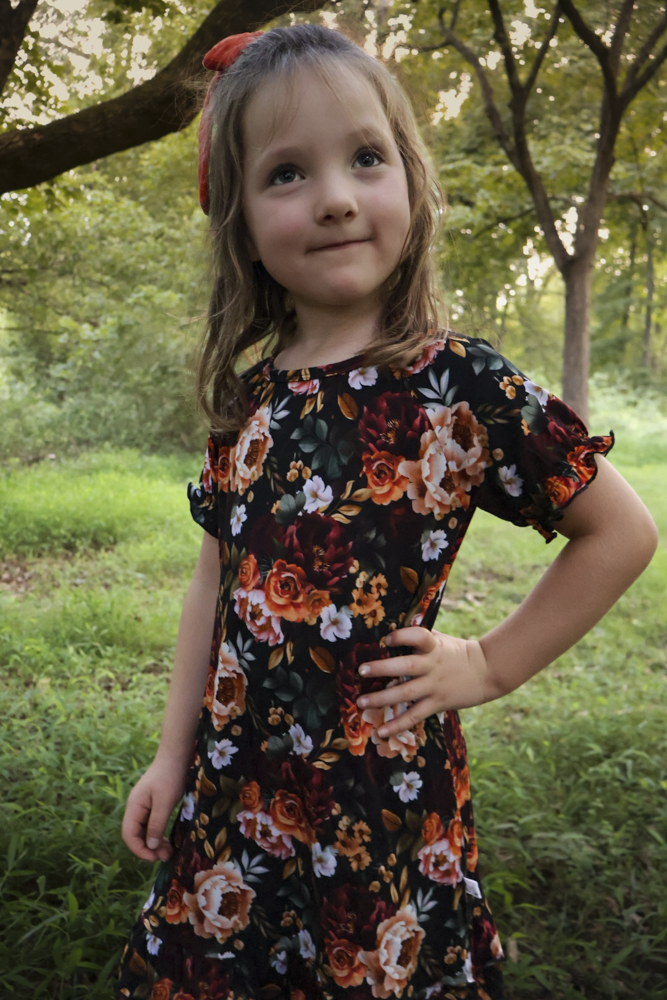 A young girl with a red hair bow stands outdoors in a grassy park area wearing a short-sleeve black nightgown with a vibrant orange, red, and white floral pattern.
