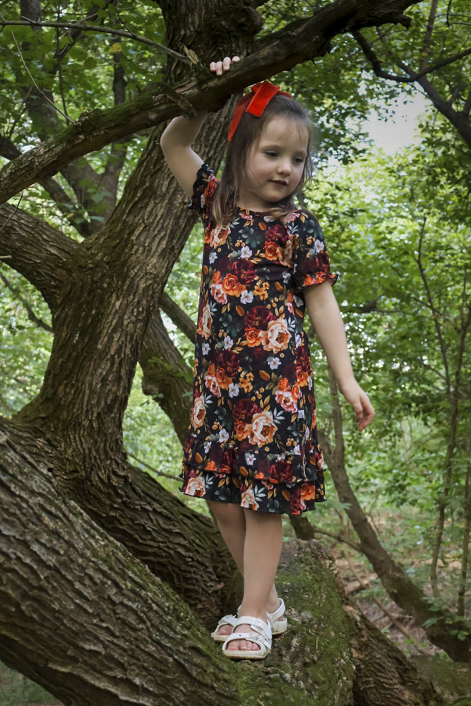 A young girl wearing a short-sleeve black floral nightgown and white sandals stands on a mossy tree trunk in a wooded outdoor setting, holding onto a branch with one hand.