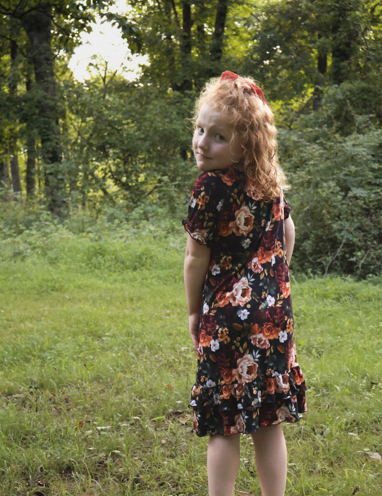 A young girl with curly red hair wearing a short-sleeve black nightgown with a vibrant orange, red, and white floral pattern, standing in a grassy park with trees in the background.