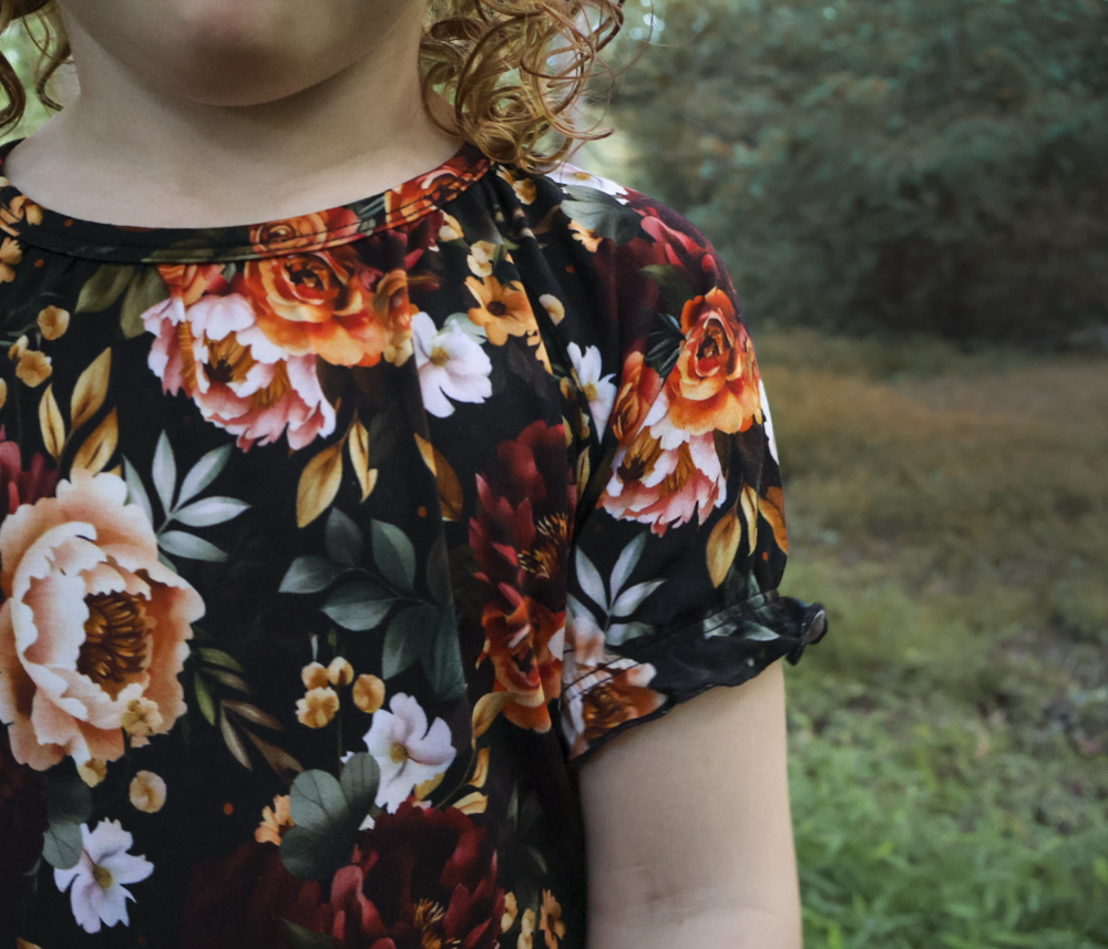 A close-up of a child wearing a short-sleeve nightgown with a black background and a large floral print featuring orange, red, pink, and white flowers with green leaves, photographed outdoors against a blurred green background.