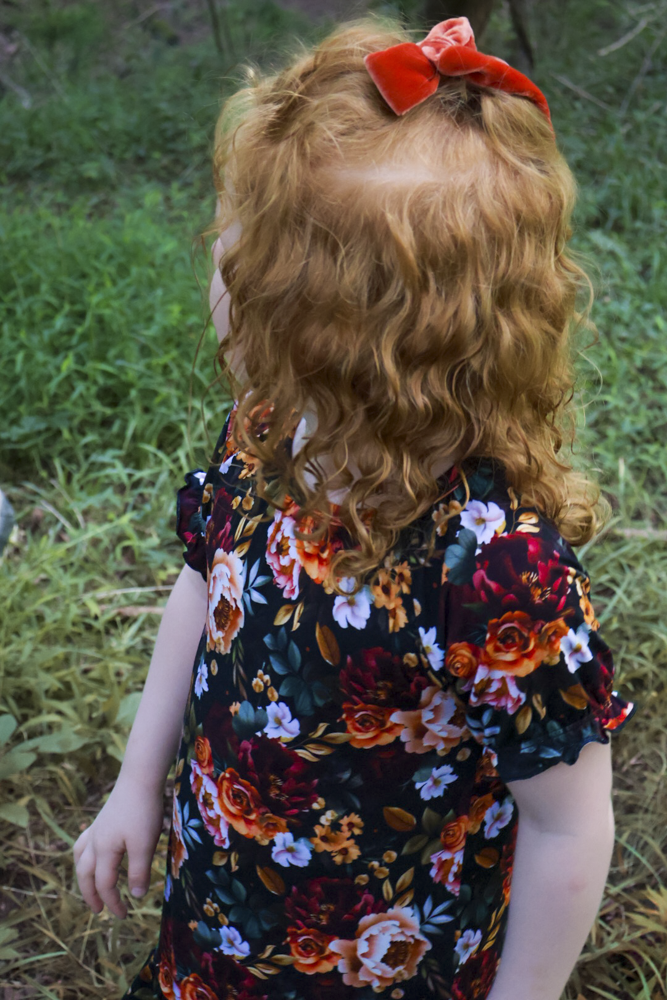A young child with curly red hair wearing a black short-sleeve nightgown with a vibrant orange, red, and white floral pattern, and a red bow in their hair, standing outdoors in a grassy area.