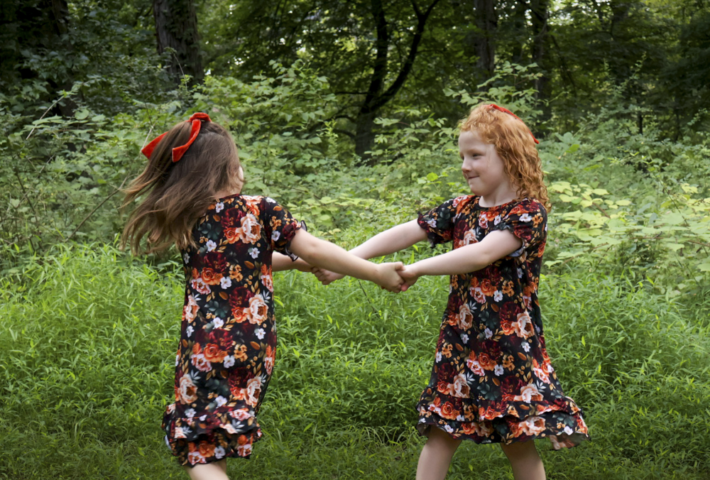 Two young girls in matching floral print nightgowns holding hands and spinning in a lush green outdoor setting.