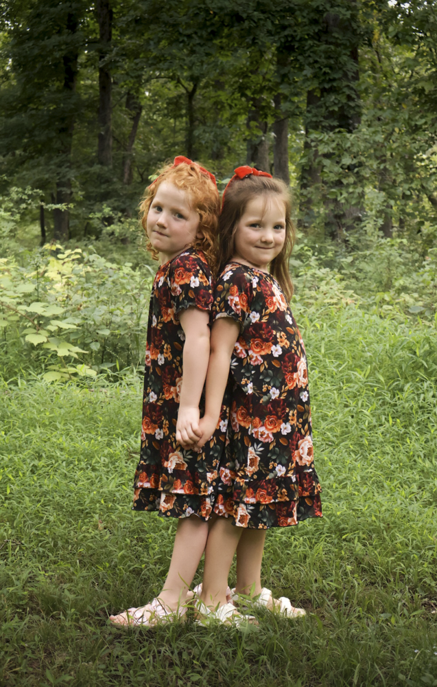 Two young girls standing back-to-back in a grassy outdoor setting, wearing matching short-sleeve floral nightgowns with a black base and red, orange, and white floral pattern, accented with red hair bows.