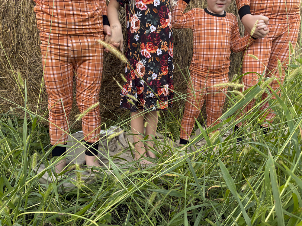 Three children standing outdoors in tall grass in front of a hay bale, wearing orange plaid matching pajamas. One child is in the center wearing a dark floral nightgown.